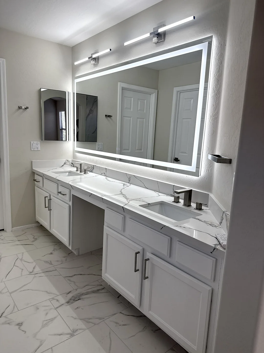 Bathroom after renovation, with a white double vanity, marble-look countertop and tile, and illuminated mirror